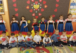 Dzieci z grupy pomarańczowej stoją na tle dekoracji z serc z przodu siedzą chłopcy ubrani w koszule z tyłu dziewczynki ubrane w suknie hiszpańskie